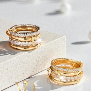 Puede incluir: Anillos de oro apilables con detalles de diamantes. Los anillos están dispuestos en pilas de cinco, con una mezcla de bandas de oro liso y bandas adornadas con pequeños diamantes brillantes. Los anillos están sobre una superficie blanca.