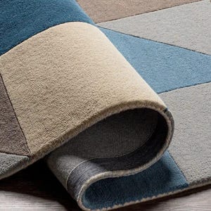 Könnte beinhalten: Aufgerollter Teppich mit geometrischem Muster in Blau-, Grau- und Beigetönen. Die Textur des Teppichs ist sichtbar, mit einer Nahaufnahme der gerollten Kante und der Unterseite.