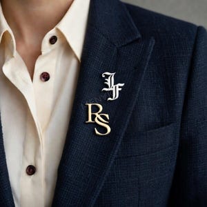 May include: Two initial pins, one in gold and one in white, are pinned on a navy blue blazer. The gold pin displays the letters "RS", while the white pin shows "JF". The blazer is worn over a cream-colored collared shirt.