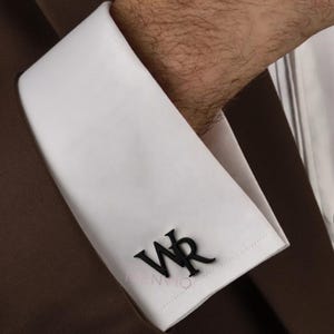 Könnte beinhalten: Nahaufnahme eines weißen Hemdärmels mit schwarzen Manschettenknöpfen. Die Manschettenknöpfe zeigen die Buchstaben "WR" in einer stilisierten Schriftart. Der Ärmel gehört zu einem braunen Anzug.
