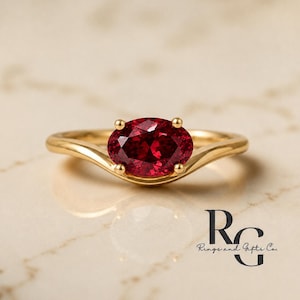 Può includere: Un anello in oro con una pietra preziosa rosso scuro dal taglio ovale fissata da quattro griffe. La fascia presenta un design liscio e curvo che si assottiglia verso l'incastonatura centrale. Il logo di Rings and Gifts Co. è visibile in basso a destra.