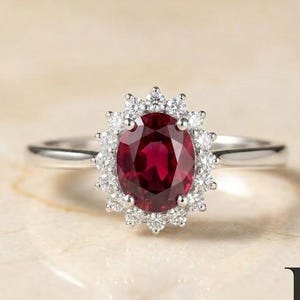 May include: A silver ring featuring a central oval-cut deep red ruby gemstone surrounded by a halo of small, clear, round-cut white diamonds. The band is polished silver, and the logo for Rings and Gifts Co. is visible in the bottom right corner.