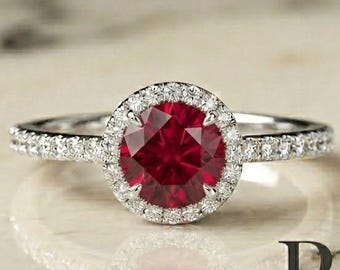 Peut inclure: Une bague argentée avec une pierre centrale ronde rouge foncé entourée d'un halo de petites pierres claires et scintillantes. L'anneau est également orné d'une rangée de petites pierres claires. Le texte "RG Rings and Gifts Co." est visible.