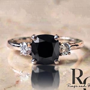 May include: A silver-toned ring featuring a large, cushion-cut black centre stone flanked by two smaller, round, clear stones. The band is polished metal. The text "RG Rings and Gifts Co." is visible in the lower right corner of the image.