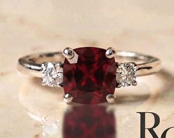 Peut inclure: Une bague argentée avec une pierre précieuse rouge foncé taille coussin au centre, entourée de deux petites pierres claires taille ronde. L'anneau est en métal poli. Le texte "RG Rings and Gifts Co." est imprimé en noir en bas à droite.
