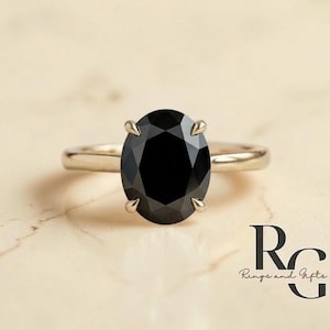 May include: A silver ring with a large, oval-cut black gemstone. The ring is set on a light-colored surface. The logo "Rings and Gifts Co." is visible in the bottom right corner.