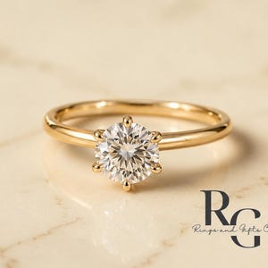 Peut inclure: Une bague en or avec une pierre pr&eacute;cieuse transparente ronde taille brillant fix&eacute;e par six griffes. L'anneau est en or lisse et poli. Le texte "RG Rings and Gifts Co." est visible dans le coin inf&eacute;rieur droit de l'image.