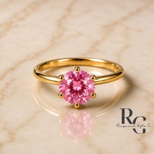 Peut inclure: Une bague en or avec une pierre pr&eacute;cieuse rose taille brillant ronde, maintenue par un sertissage &agrave; six griffes. L'anneau est en or lisse et poli. Le logo de Rings and Gifts Co. est visible dans le coin inf&eacute;rieur droit.