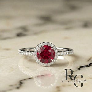 May include: A silver-toned ring featuring a round, deep red centre stone surrounded by a halo of small, clear, sparkling stones. The band is also set with a row of small, clear stones. The text "RG Rings and Gifts Co." is visible in the corner.
