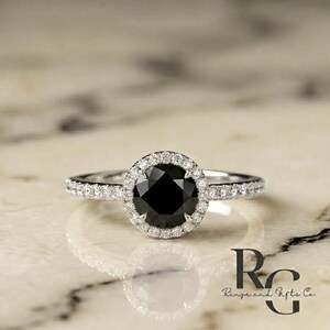 May include: A silver-toned ring featuring a large, round, faceted black centre stone surrounded by a halo of small, clear, sparkling stones. The band is also set with a row of small, clear stones along each side. The text RG Rings and Gifts Co. is visible.