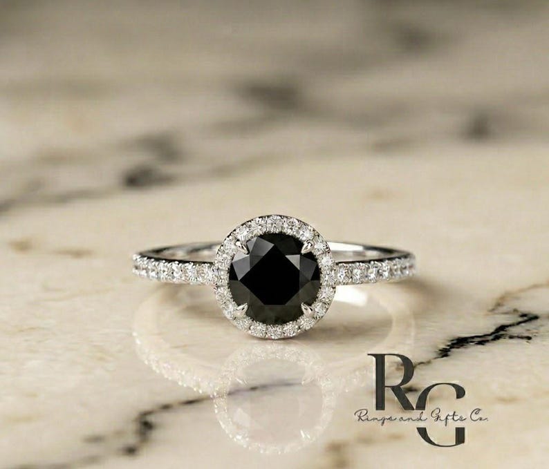 May include: A silver-toned ring featuring a large, round, faceted black centre stone surrounded by a halo of small, clear, sparkling stones. The band is also set with a row of small, clear stones along each side. The text RG Rings and Gifts Co. is visible.