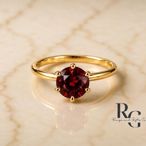 Peut inclure: Une bague en or avec une pierre pr&eacute;cieuse ronde rouge fonc&eacute; mont&eacute;e sur un sertissage &agrave; six griffes. L'anneau est en or lisse et poli. Le texte RG Rings and Gifts Co. est visible dans le coin inf&eacute;rieur droit, indiquant la marque.