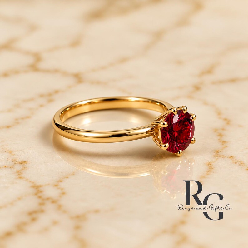 Peut inclure: Une bague en or avec une pierre pr&eacute;cieuse rouge fonc&eacute;, ronde et &agrave; facettes, maintenue par un sertissage &agrave; six griffes. L'anneau est en or lisse et poli. Le logo de Rings and Gifts Co. est visible dans le coin inf&eacute;rieur droit.