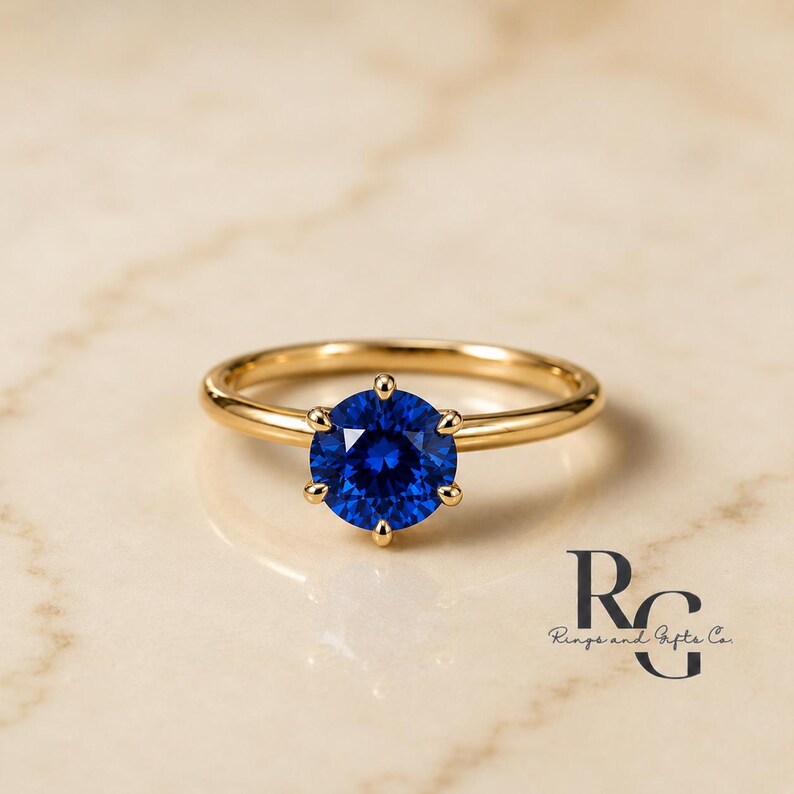 Peut inclure: Une bague en or avec une pierre pr&eacute;cieuse ronde d'un bleu profond, taill&eacute;e en brillant et fix&eacute;e par six griffes. L'anneau est en or lisse et poli. Le texte "RG Rings and Gifts Co." est visible dans le coin inf&eacute;rieur droit de l'image.