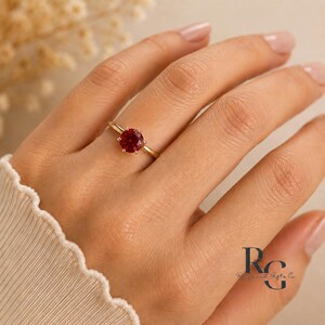 Peut inclure: Une bague en or avec une pierre pr&eacute;cieuse rouge fonc&eacute;, ronde et facett&eacute;e, mont&eacute;e sur un sertissage &agrave; six griffes. L'anneau est fin et en or poli. Le logo RG est visible en bas &agrave; droite, avec le texte Reign and Gifts Co. en dessous.