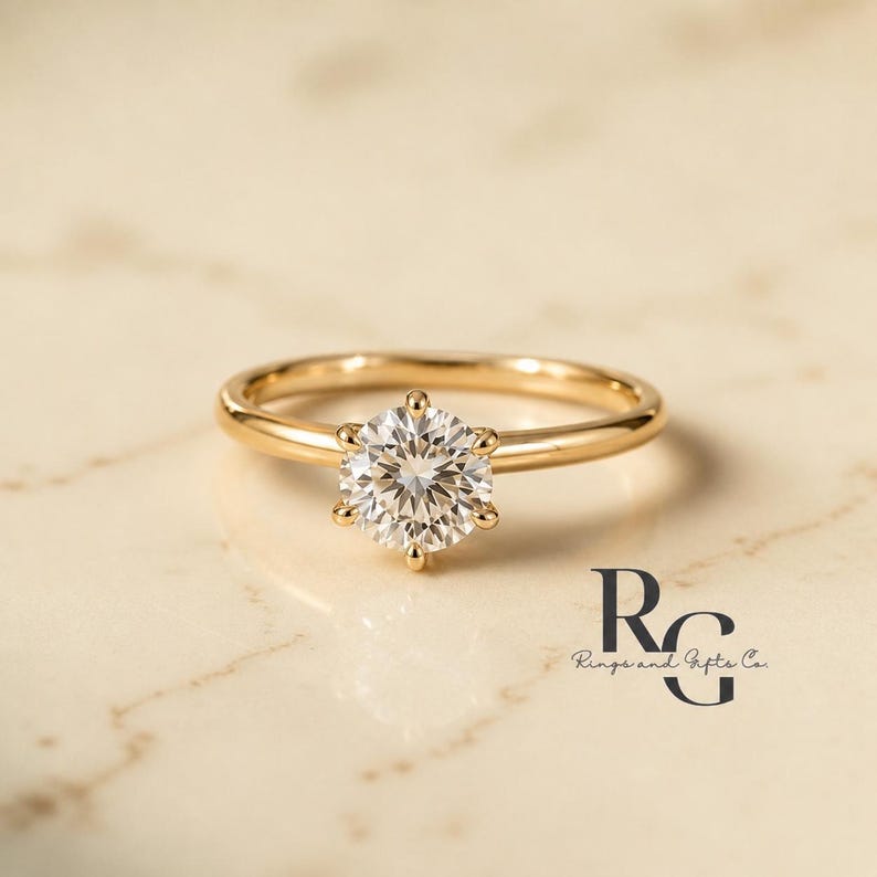Peut inclure: Une bague en or avec une pierre pr&eacute;cieuse transparente ronde taille brillant fix&eacute;e par six griffes. L'anneau est en or lisse et poli. Le texte "RG Rings and Gifts Co." est visible dans le coin inf&eacute;rieur droit de l'image.