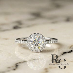 May include: A silver-coloured ring featuring a large, round, brilliant-cut centre stone surrounded by a halo of smaller pav&eacute;-set stones. The slender band is also adorned with a row of tiny, sparkling stones along each side of the central setting.