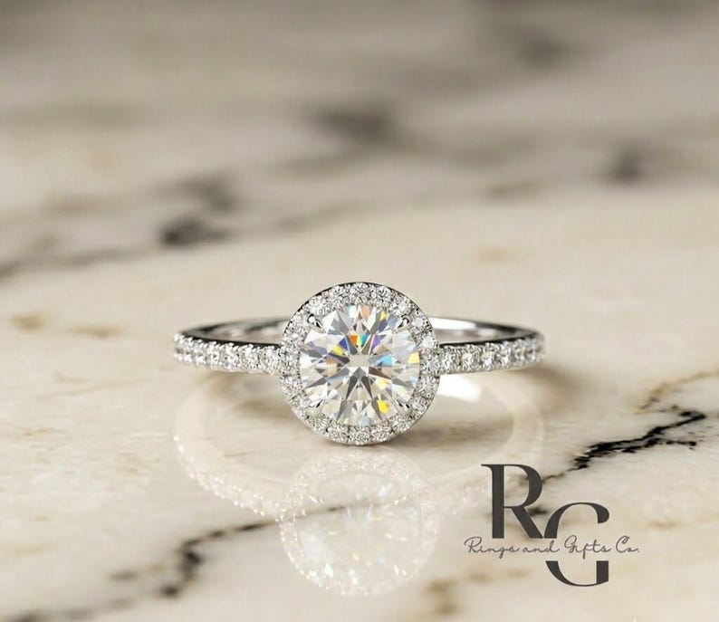 May include: A silver-coloured ring featuring a large, round, brilliant-cut centre stone surrounded by a halo of smaller pav&eacute;-set stones. The slender band is also adorned with a row of tiny, sparkling stones along each side of the central setting.