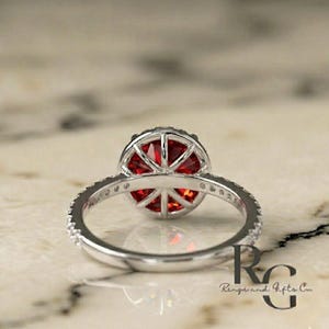 May include: A silver ring featuring a round, faceted red gemstone set in a basket-style mounting. The band is adorned with small, clear stones along the sides. The text RG Rings and Gifts Co is visible in the lower right corner of the image.