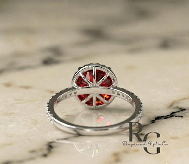 May include: A silver ring featuring a round, faceted red gemstone set in a basket-style mounting. The band is adorned with small, clear stones along the sides. The text RG Rings and Gifts Co is visible in the lower right corner of the image.