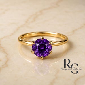 Peut inclure: Une bague en or avec une pierre pr&eacute;cieuse ronde, facett&eacute;e et violette fonc&eacute;e, maintenue par six griffes. L'anneau est en or lisse et poli. Le texte "RG Rings and Gifts Co." est visible dans le coin inf&eacute;rieur droit, &eacute;crit en noir.