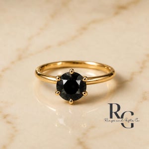 Peut inclure: Une bague en or avec une pierre noire ronde &agrave; facettes mont&eacute;e sur un sertissage &agrave; six griffes. L'anneau est en or lisse et poli. Le texte RG Rings and Gifts Co. est visible dans le coin inf&eacute;rieur droit, &eacute;crit dans une police serif noire.