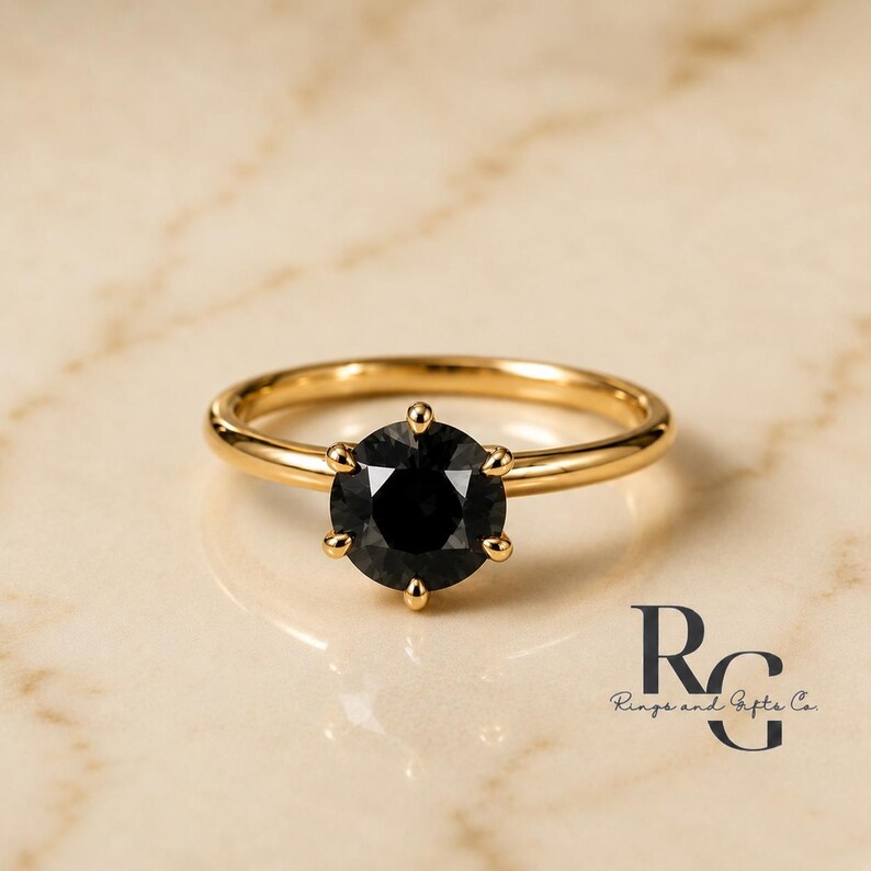 Peut inclure: Une bague en or avec une pierre noire ronde &agrave; facettes mont&eacute;e sur un sertissage &agrave; six griffes. L'anneau est en or lisse et poli. Le texte RG Rings and Gifts Co. est visible dans le coin inf&eacute;rieur droit, &eacute;crit dans une police serif noire.