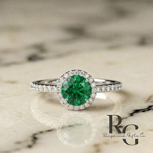 May include: A silver-toned ring featuring a round, vibrant green centre stone surrounded by a halo of small, clear, sparkling stones. The band is also set with a row of tiny, clear stones along each side, creating a bright, reflective appearance.