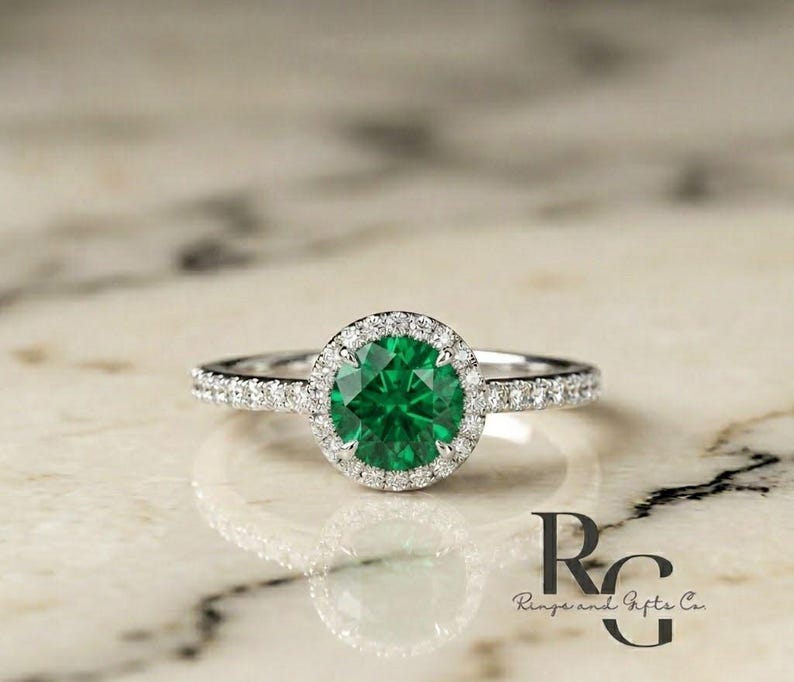May include: A silver-toned ring featuring a round, vibrant green centre stone surrounded by a halo of small, clear, sparkling stones. The band is also set with a row of tiny, clear stones along each side, creating a bright, reflective appearance.