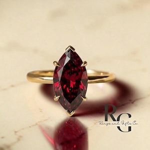 May include: A gold ring featuring a marquise-cut, deep red gemstone. The gemstone is set with four prongs, and the band is a simple, polished gold. The ring is displayed on a light-colored surface.