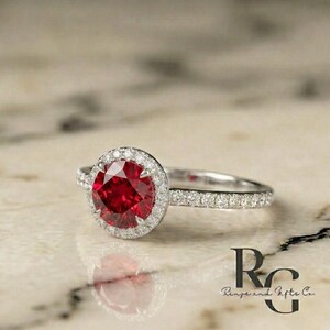 May include: A silver-toned ring featuring a large, round, faceted red centre stone surrounded by a halo of small, clear, sparkling stones. The band is also encrusted with small, clear stones. The text "RG Rings and Gifts Co." is visible.