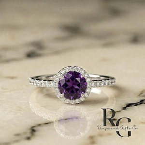 May include: A silver-toned ring featuring a round, deep purple amethyst centre stone surrounded by a halo of small, clear, sparkling stones. The band is also set with a row of small, clear stones along each side, with the text Rings and Gifts Co.