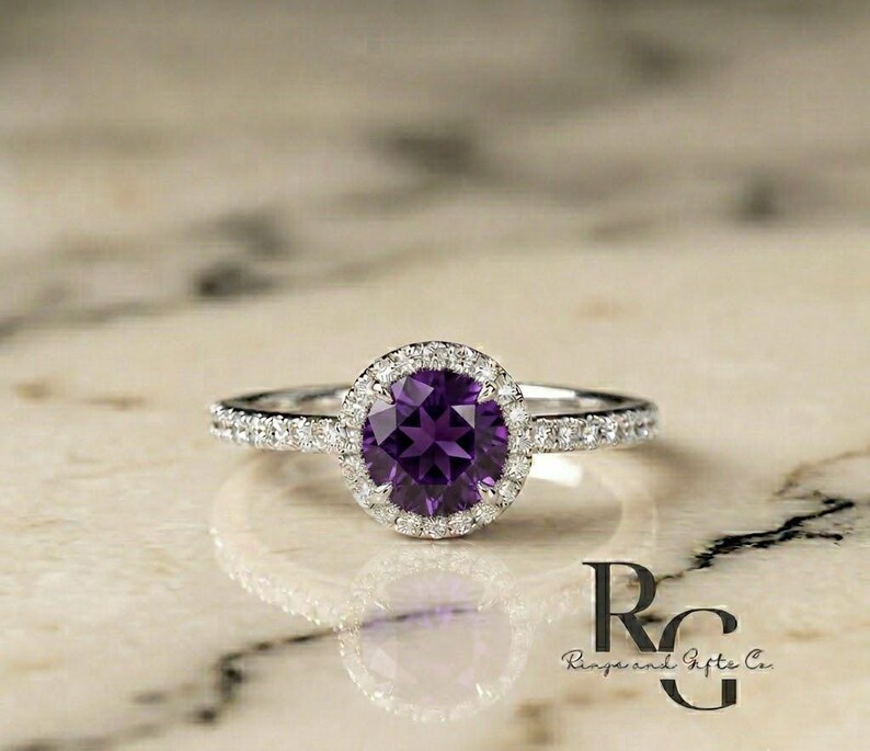 May include: A silver-toned ring featuring a round, deep purple amethyst centre stone surrounded by a halo of small, clear, sparkling stones. The band is also set with a row of small, clear stones along each side, with the text Rings and Gifts Co.