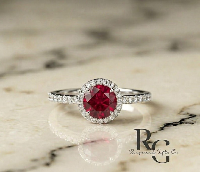 May include: A silver-toned ring featuring a round, deep red centre stone surrounded by a halo of small, clear, sparkling stones. The band is also set with a row of small, clear stones. The text "RG Rings and Gifts Co." is visible in the corner.