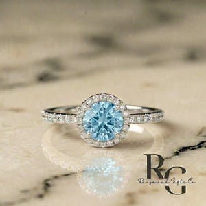 May include: A silver-coloured ring featuring a round, light blue centre stone surrounded by a halo of small, clear, sparkling stones. The band is also set with small, clear stones along the sides. The text "RG Rings and Gifts Co." is visible.