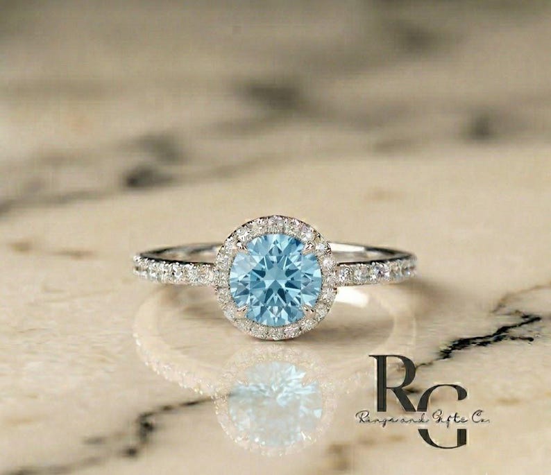 May include: A silver-coloured ring featuring a round, light blue centre stone surrounded by a halo of small, clear, sparkling stones. The band is also set with small, clear stones along the sides. The text "RG Rings and Gifts Co." is visible.