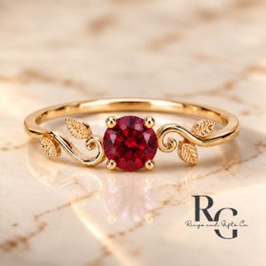 Peut inclure: Une bague en or ornée d'une pierre précieuse ronde rouge foncé au centre. L'anneau est conçu avec de délicats motifs de feuilles dorées polies s'étendant des deux côtés de la pierre centrale. Le texte Rings and Gifts Co. est visible.