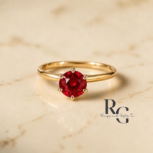 Peut inclure: Une bague en or avec une pierre pr&eacute;cieuse rouge vif, ronde et &agrave; facettes, mont&eacute;e sur un sertissage &agrave; six griffes. L'anneau est lisse et poli. Le texte RG Rings and Gifts Co. est visible dans le coin inf&eacute;rieur droit de l'image.