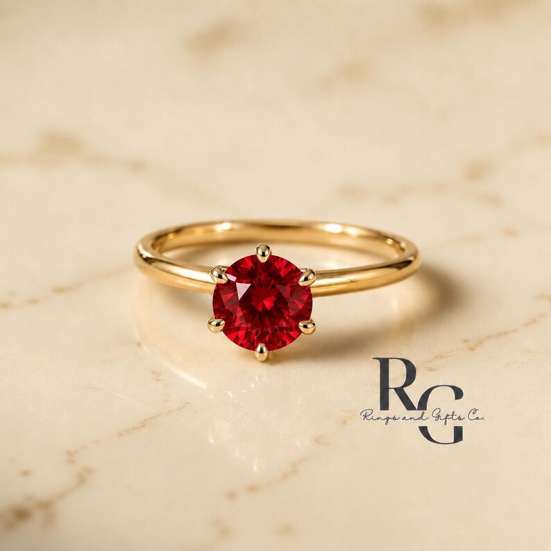 Peut inclure: Une bague en or avec une pierre pr&eacute;cieuse rouge vif, ronde et &agrave; facettes, mont&eacute;e sur un sertissage &agrave; six griffes. L'anneau est lisse et poli. Le texte RG Rings and Gifts Co. est visible dans le coin inf&eacute;rieur droit de l'image.