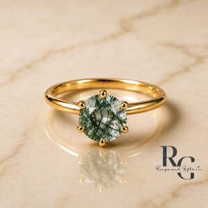 Peut inclure: Une bague en or avec une pierre pr&eacute;cieuse d'agate mousse ronde et facett&eacute;e aux inclusions vert fonc&eacute;. La pierre est fix&eacute;e par six griffes sur un anneau en or poli. Le texte RG Rings and Gifts Co. est visible dans le coin inf&eacute;rieur droit.