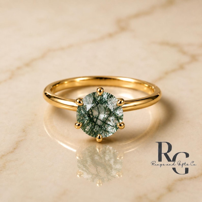 Peut inclure: Une bague en or avec une pierre pr&eacute;cieuse d'agate mousse ronde et facett&eacute;e aux inclusions vert fonc&eacute;. La pierre est fix&eacute;e par six griffes sur un anneau en or poli. Le texte RG Rings and Gifts Co. est visible dans le coin inf&eacute;rieur droit.
