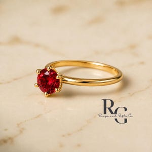 Peut inclure: Une bague en or avec une pierre pr&eacute;cieuse rouge vif, ronde et &agrave; facettes, maintenue par un sertissage &agrave; six griffes. L'anneau est en or lisse et poli. Le logo de Rings and Gifts Co. est visible dans le coin inf&eacute;rieur droit de l'image.