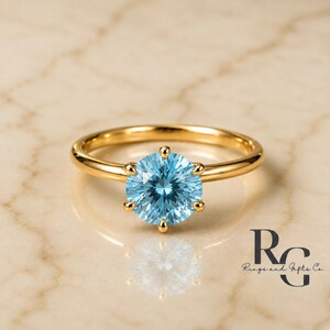 Peut inclure: Une bague en or avec une pierre pr&eacute;cieuse ronde bleu clair &agrave; facettes, fix&eacute;e par un sertissage &agrave; six griffes. L'anneau est en or lisse et poli. Le texte "RG Rings and Gifts Co." est visible dans le coin inf&eacute;rieur droit de l'image.