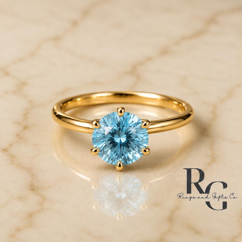 Peut inclure: Une bague en or avec une pierre pr&eacute;cieuse ronde bleu clair &agrave; facettes, fix&eacute;e par un sertissage &agrave; six griffes. L'anneau est en or lisse et poli. Le texte "RG Rings and Gifts Co." est visible dans le coin inf&eacute;rieur droit de l'image.