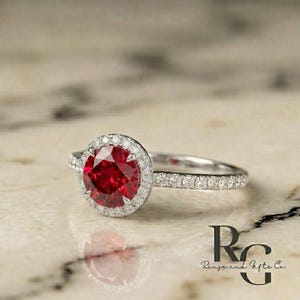 May include: A silver-toned ring featuring a large, round, faceted red centre stone surrounded by a halo of small, clear, sparkling stones. The band is also encrusted with small, clear stones. The text "RG Rings and Gifts Co." is visible.