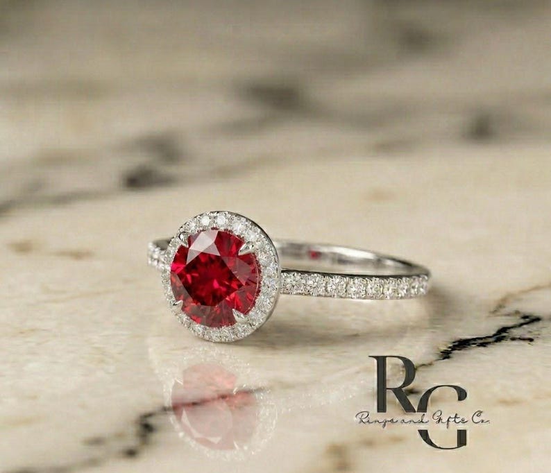 May include: A silver-toned ring featuring a large, round, faceted red centre stone surrounded by a halo of small, clear, sparkling stones. The band is also encrusted with small, clear stones. The text "RG Rings and Gifts Co." is visible.