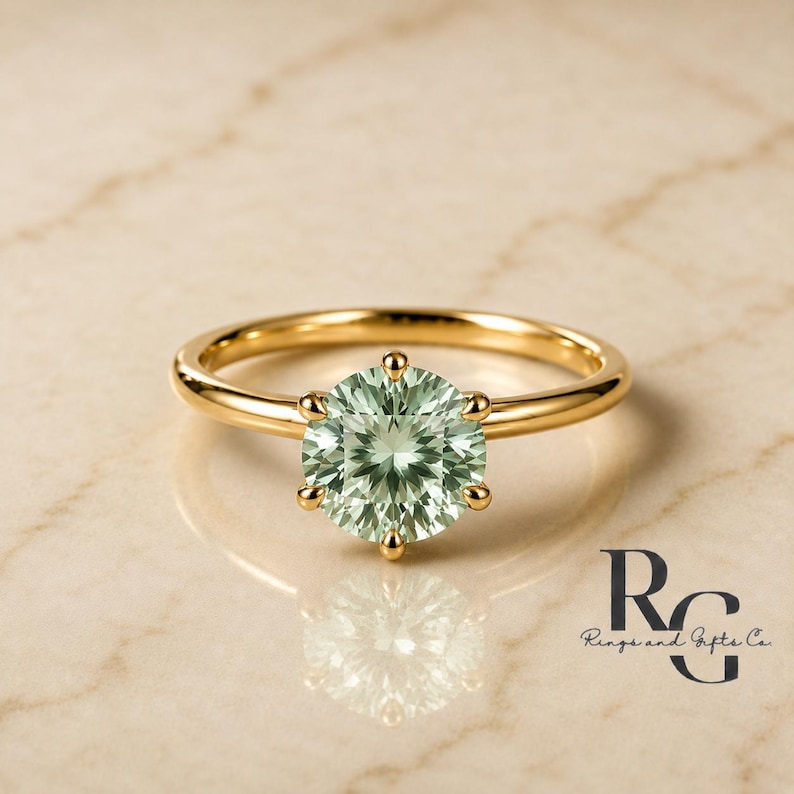 Peut inclure: Une bague en or avec une pierre pr&eacute;cieuse ronde, facett&eacute;e et vert clair, maintenue par un sertissage &agrave; six griffes. L'anneau est en or lisse et poli. Le texte "RG Rings and Gifts Co." est visible dans le coin inf&eacute;rieur droit.