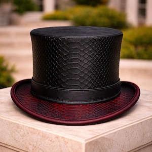 Può includere: Un cappello a cilindro nero con un motivo a pelle di serpente sui lati e un motivo a pelle di serpente bordeaux sulla tesa. Il cappello ha una fascia nera al centro. Accessorio formale, su una superficie di pietra.