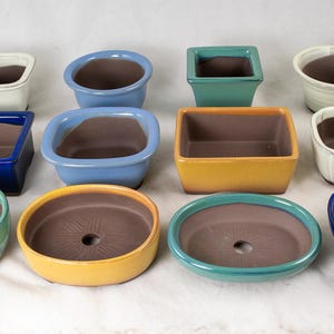 May include: A collection of ceramic bonsai pots in various shapes and colors, including blue, green, yellow, and white. The pots have a brown interior and are designed for planting small trees or plants. The collection includes square, oval, and round designs.