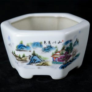 May include: White ceramic planter with a hexagonal shape and a painted landscape scene. The artwork depicts mountains, trees, and a building with text. The planter has a glossy finish and is suitable for small plants.